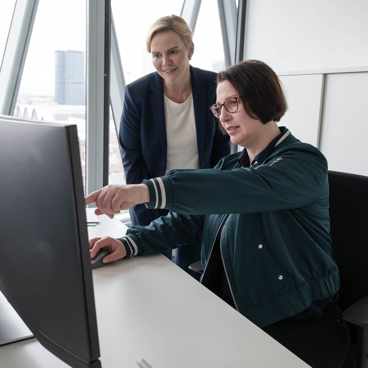 Zwei NHW-Mitarbeiterinnen schauen lächelnd in einen PC-Monitor, während die eine sitzt und die andere ihr über die Schulter schaut. Sie befinden sich in einem Büro im Westhafen Tower in Frankfurt.