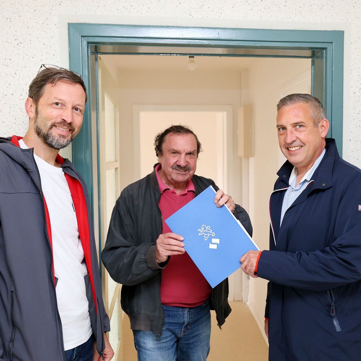 Ein älterer Herr steht zwischen einem NHW Mitarbeiter sowie dem Geschäftsführer der Evangelischen Wohnraumhilfe in der Tür seiner neuen Wohnung und bekommt Unterlagen ausgehändigt. Alle gucken lächelnd in die Kamera.