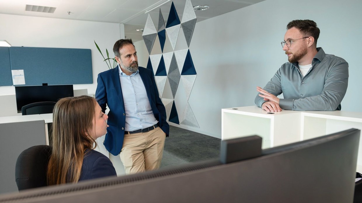Drei NHW-Mitarbeiter sitzen bzw. stehen in einem Büro mit vielen Rechnern auf den Schreibtischen im Westhafen Tower in Frankfurt und führen ein konzentriertes Arbeitsgespräch.