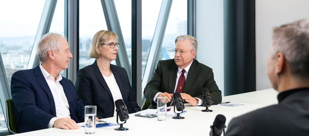 Von links nach rechts ist Herr Dr. Hain, Frau Fontaine-Kretschmer und Herr Dr. Westphal auf dem Bild zusehen. Sie sitzen zusammen an einem Konferenz-Tisch und werden von einem Interviewer, der im Anschnitt des Bildes ist, interviewt.