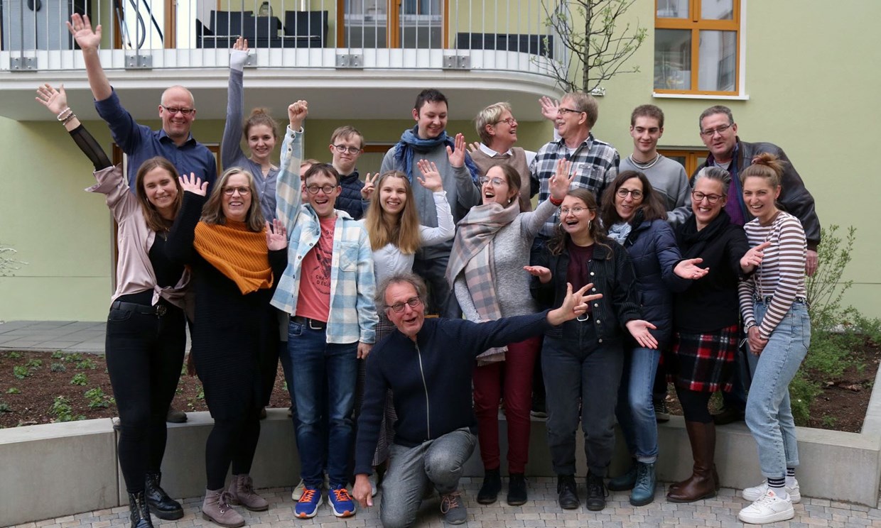 Gruppenfoto von der inklusiven Wohngemeinschaft in der Freiligrathstraße in Frankfurt. Sie winken vor dem Gebäude in die Kamera.