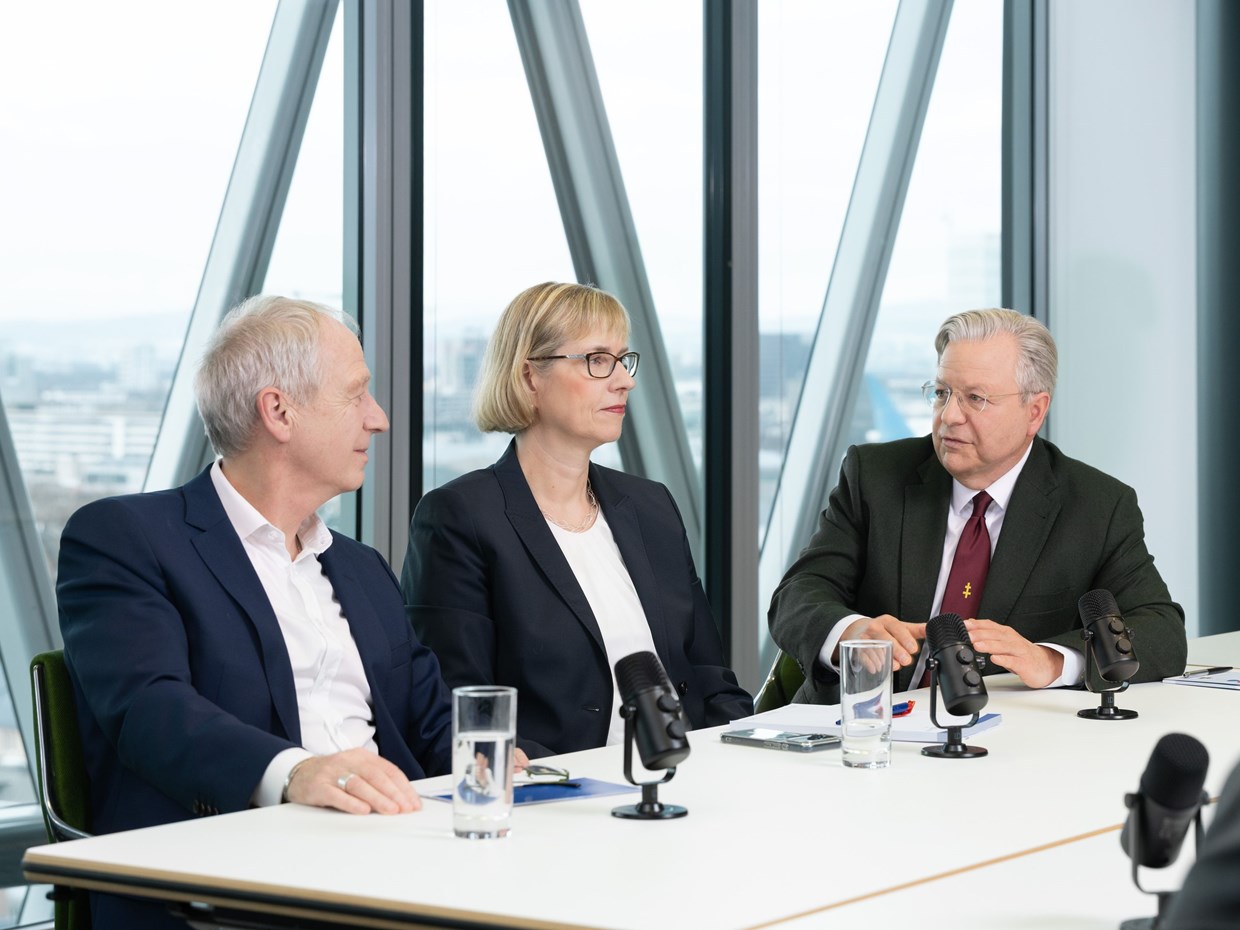 Von links nach rechts ist Herr Dr. Hain, Frau Fontaine-Kretschmer und Herr Dr. Westphal auf dem Bild zusehen. Sie sitzen zusammen an einem Konferenz-Tisch und werden von einem Interviewer, der im Anschnitt des Bildes ist, interviewt.