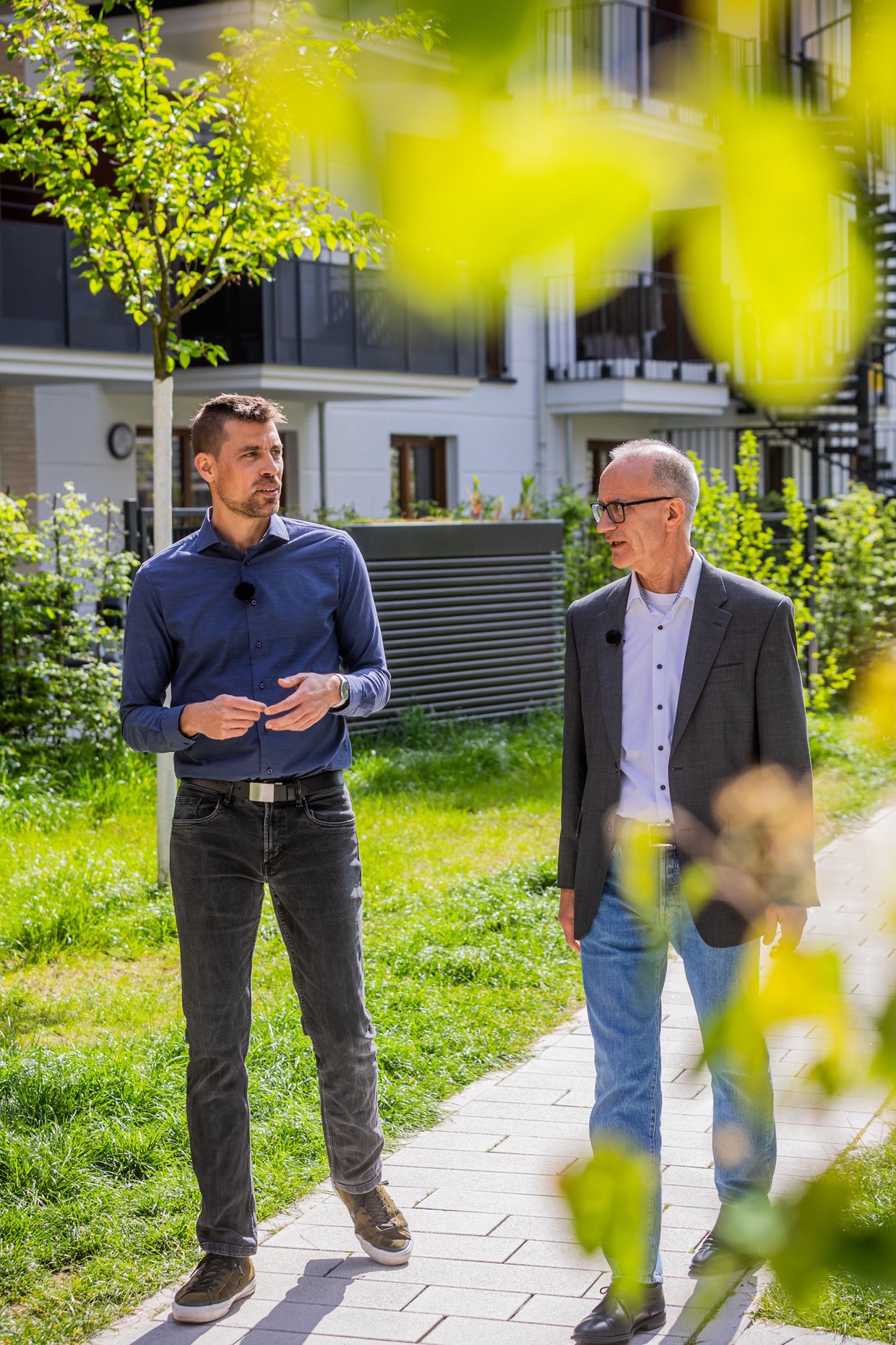 Zwei Mitarbeiter der NHW spazieren durch Schönhof-Viertel in Frankfurt. Ansicht durch grüne Blätter.