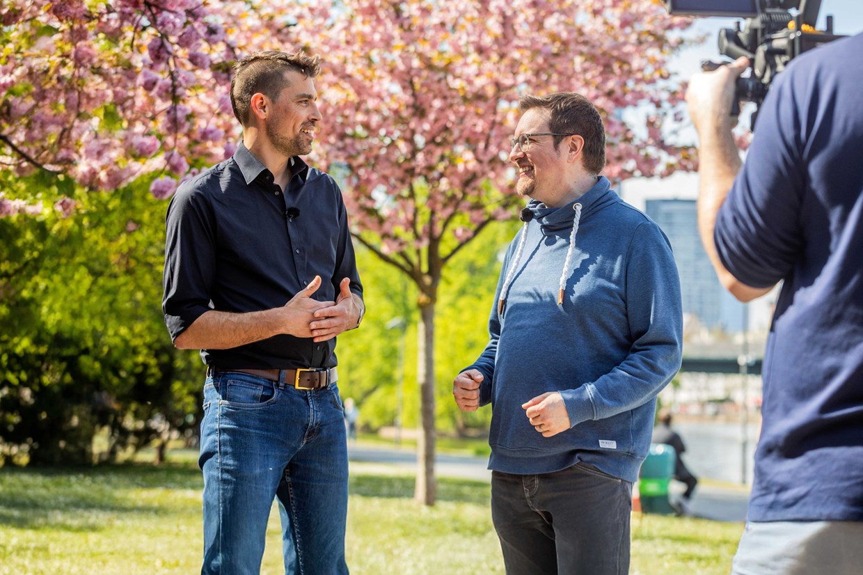 Zwei NHW Mitarbeiter unterhalten sich am sonnigen Mainufer und werden dabei von einer Filmcrew begleitet.