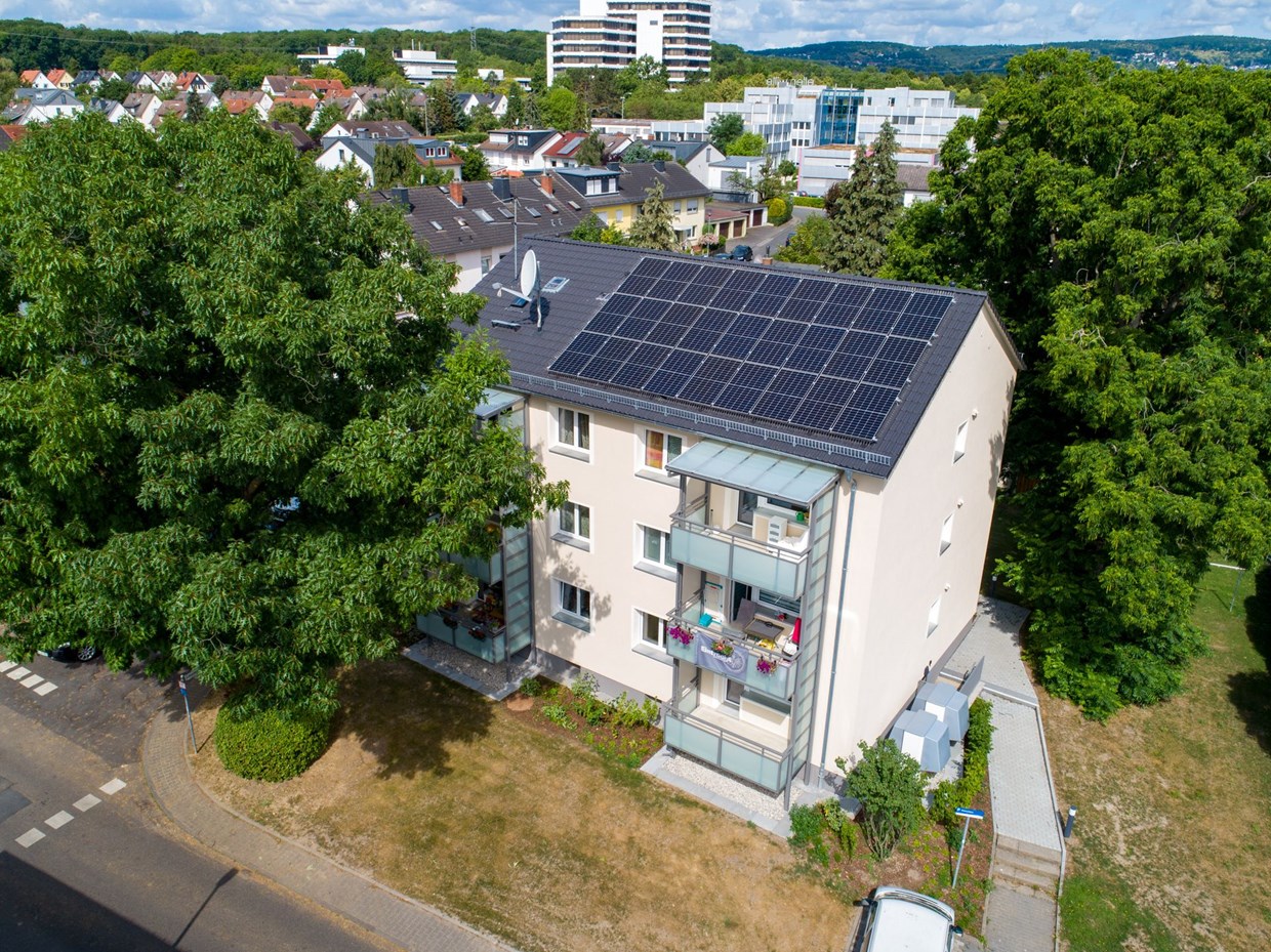 Hintergrundbild: Blick von oben auf ein Mietshaus mit Solar-Panelen auf dem Dach. Dahinter sind weitere Wohnhäuser sowie Bäume zu sehen.